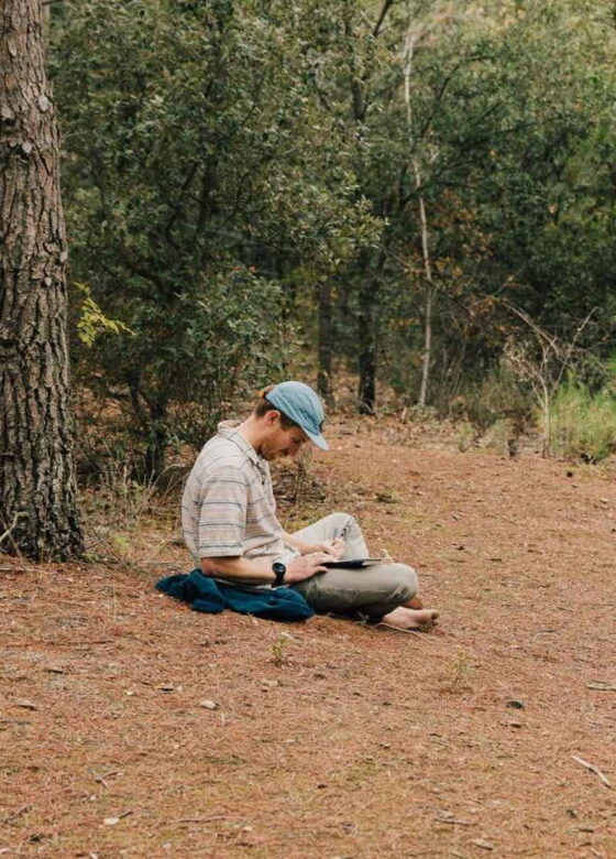 A man sitting cross legged on the forest floor reading